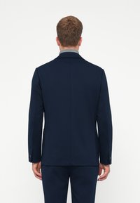 Homem em pé com as costas voltadas para a câmara, vestindo um blazer escuro azul-marinho e calças combinando, contra um fundo branco liso.