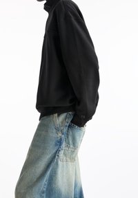 Felpa nera con zip e colletto alto, realizzata in tessuto morbido. Indossata con jeans azzurri larghi dotati di tasca a toppa.