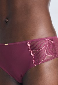 Burgundy bikini bottom with mesh panel featuring pink floral embroidery, scalloped edges, and gold hardware detail. Smooth, stretchy fabric.