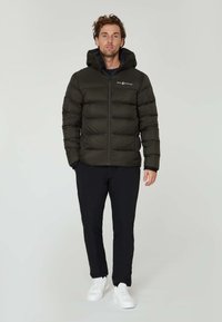 Sort pufferjakke med hætte, der har quiltet tekstur og frontlynlås. Kombineret med sorte bukser og hvide sneakers, stående mod en neutral baggrund.