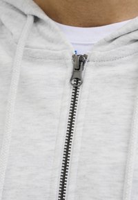 Sudadera con cremallera de color gris claro, hecha de una tela suave, que presenta una cremallera y cordones negros, con una textura acanalada cerca del escote.