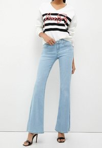 Pull blanc à rayures horizontales noires avec lettres rouges, associé à un jean évasé bleu clair et des talons ouverts noirs.