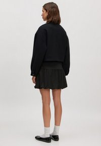 Zwarte cropped sweatshirt met verlaagde schouders, gecombineerd met een geplooide zwarte minirok. Model draagt zwarte ballerina's en witte sokken. Minimalistisch ontwerp.