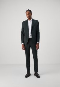 Dark green suit with a slim fit, single-breasted jacket and matching trousers. White dress shirt underneath, worn with black loafers.