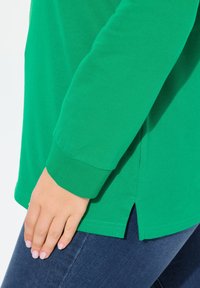 Maglietta verde in cotone a maniche lunghe con polsini a costine, fessure laterali all'orlo e una texture liscia, abbinata a jeans blu scuro.