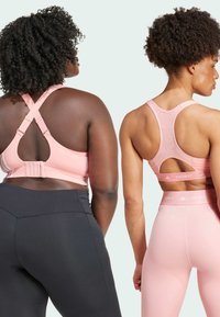 Dos personas llevan sujetadores deportivos rosas con tirantes ajustables, que presentan un diseño de espalda abierta y una banda TECHFIT. A la izquierda, lleva leggings negros; a la derecha, leggings rosas.
