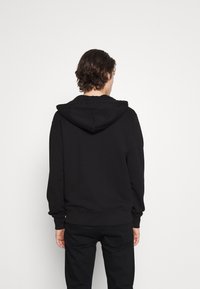 Sudadera de algodón negra con una textura suave, capucha con cordones ajustables y puños y cintura acanalados. Diseño liso, sin logotipos ni estampados visibles.