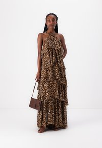 Maxi haljina s leopard uzorkom, slojevima volana, halter izrezom i prozirnom teksturom. Model drži smeđu torbu za rame, stoji na jednoličnoj pozadini.