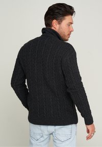 Dunkelgrauer Kabelstrickpullover mit hohem Kragen und geripptem Saum, ausgestattet mit strukturiertem Stoff und langen Ärmeln. Getragen mit hellen Jeans.