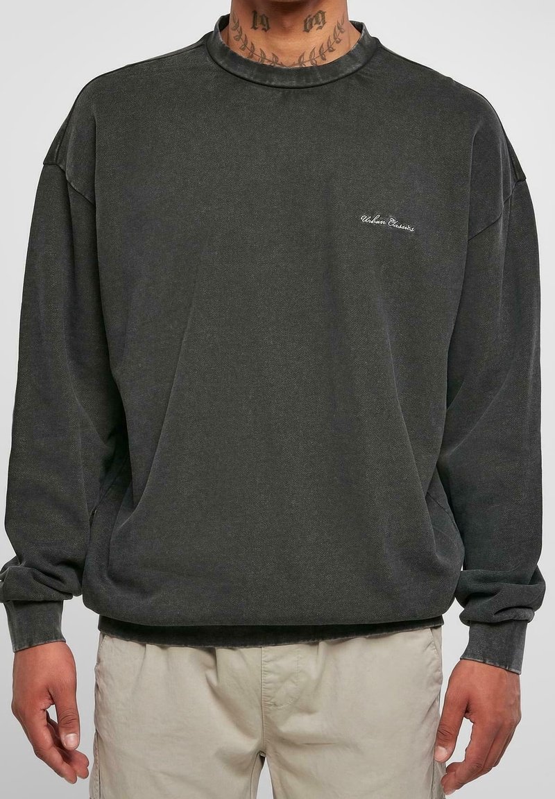Homme portant un sweat-shirt gris foncé à manches longues avec un petit texte blanc sur la poitrine et un pantalon beige clair sur un fond uni.