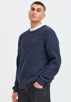Navy blue knitted sweater with a crew neckline, long sleeves, and ribbed cuffs; textured fabric with a loose fit, paired with black jeans.