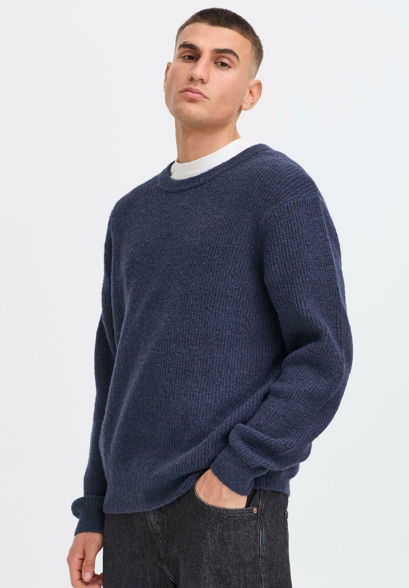 Pull en maille bleu marine avec un col rond, des manches longues et des poignets côtelés ; tissu texturé avec une coupe ample, associé à un jean noir.