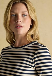 Prúžkovaný rebrovaný top v krémovej a námorníckej farbe, s okrúhlym výstrihom a priliehavým strihom. Model má dlhé, vlnité blond vlasy a neutrálne pozadie.