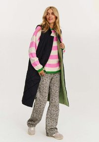 gepolsterte schwarze Weste, mehrfarbig gestreifter Pullover mit rosa und grünen Akzenten, Hose im Leopardenmuster und graue Slipper.