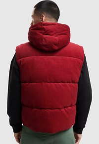 Rød cordfløyels puffer vest med hette. Har vertikale ribbete teksturer, quiltet design, og en avslappet passform. Båret over en svart langermet skjorte.