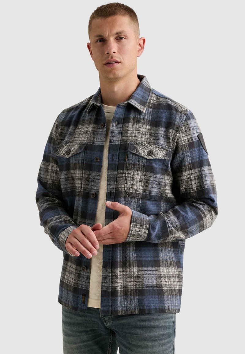 Homme portant une chemise à carreaux bleue, noire et blanche par-dessus un haut beige, debout, les mains jointes devant lui.