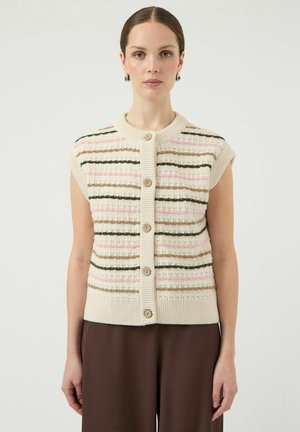 Mujer con chaleco de punto sin mangas color crema con botones y franjas horizontales en rosa, verde y beige, combinada con pantalones marrones, de pie frente a un fondo blanco.