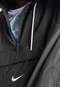 Nike lättviktsanorak i mörkgrön, med en texturerad yta, halvlång dragkedja, vit dragsko och broderad logotypdetalj.