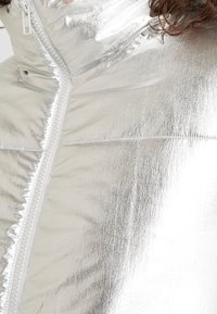 Sølv puffjakke med en skinnende, reflekterende overflade, der har en frontlynlås og stikninger på materialet.