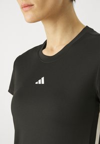 Musta urheilupaita, jossa on pyöreä pääntie, lyhyet hihat ja pieni valkoinen logo rintakehässä. Tasainen rakenne.