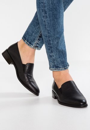 Zapatos negros de cuero de tacón bajo, tipo slip-on, llevados con unos vaqueros de mezclilla azul con un dobladillo ligeramente vuelto sobre un suelo blanco.