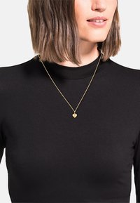 Collana con ciondolo a forma di cuore in oro su una catena sottile, indossata sopra una maglietta nera a maniche lunghe, caratterizzata da una superficie liscia e un design semplice.