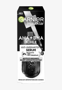 Die Verpackung des Garnier Hautklar AHA + BHA Serums zeigt eine schwarz-weiße Box mit einer transparenten Tropfflasche, die die aktiven Inhaltsstoffe in fettem Text hervorhebt.