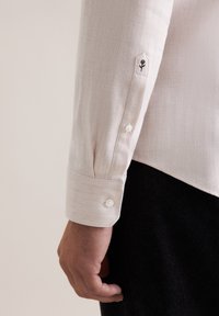 Camicia herringbone beige chiaro con polsino a bottone. Presenta un singolo dettaglio floreale ricamato vicino al polsino e bottoni bianchi.