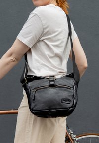 Schwarze Leder-Crossbody-Tasche mit strukturiertem Finish, vorderer Reißverschlusstasche und verstellbarem Tragegurt, auf der Schulter getragen vor einem grauen Hintergrund.