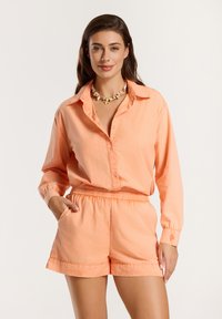 Camicia button-up pesca con maniche lunghe, abbinata a pantaloni corti a taglio rilassato dello stesso colore. Entrambi gli articoli presentano una texture morbida e una vita elastica.
