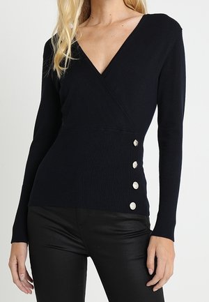 Femme portant un haut noir à manches longues en V avec quatre boutons argentés sur le côté, associé à un pantalon noir.