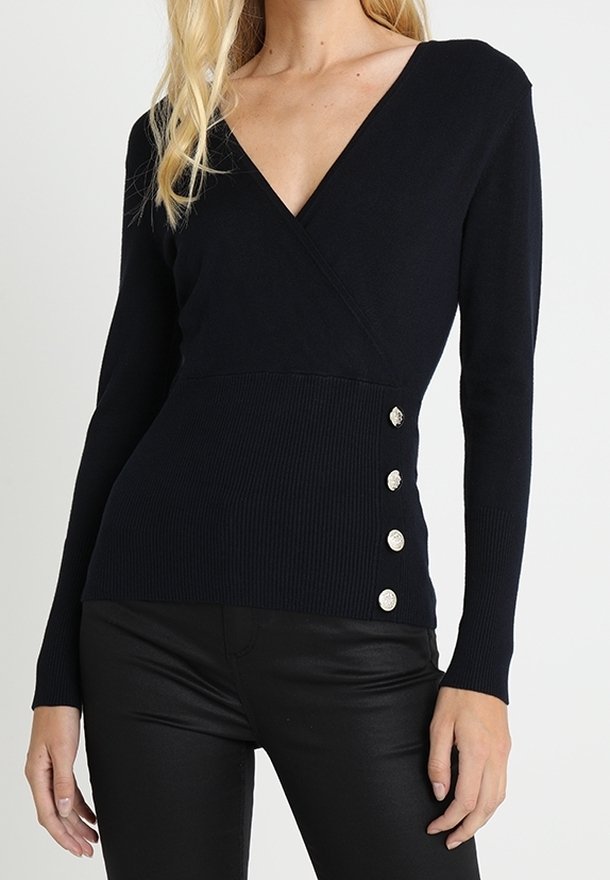 Femme portant un haut noir à manches longues en V avec quatre boutons argentés sur le côté, associé à un pantalon noir.
