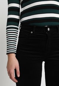 Arm en torso dragen een groen, wit en zwart gestreept langslavop T-shirt, ingestopt in hoge zwarte broek met een knoop en rits.
