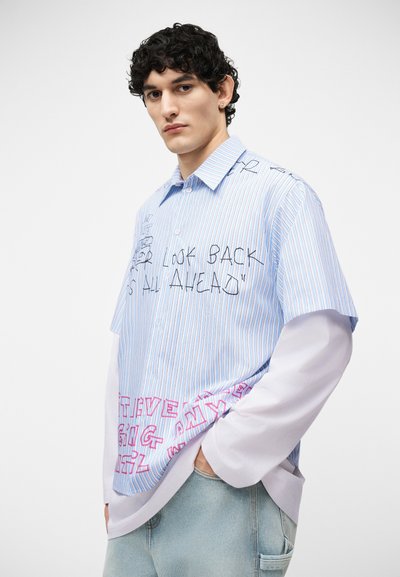 Jeune homme aux cheveux bouclés portant une chemise à rayures bleu clair superposée avec du texte manuscrit noir et rose, et un jean en denim clair.
