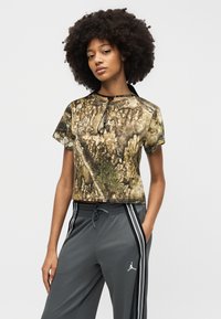 Νεαρή γυναίκα με σγουρά μαλλιά, που φοράει ένα καφέ crop top με παραλλαγή και γκρι παντελόνι με κορδόνι, ριγέ στο πλάι και λογότυπο.