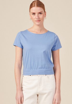 Top bleu clair à manches courtes en tissu doux. Dispose d'un col rond et d'une taille cintrée. Porté avec un pantalon de couleur claire.