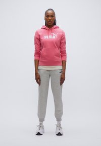 Φούξια φούτερ με κουκούλα και λευκό λογότυπο, γκρι φόρμα και λευκά αθλητικά παπούτσια. Το μοντέλο στέκεται μπροστά σε απλό φόντο, με τα χέρια χαλαρά στο πλάι.