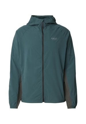 Mand iført teal og grå fuld-zip hættetrøje og matchende bukser, stående med hænderne i lommerne mod en ensfarvet baggrund.