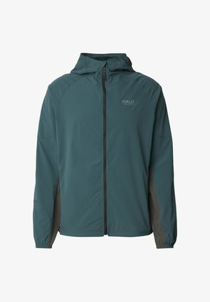Man draagt een turquoise en grijze full-zip capuchonjas en bijpassende broek, staat met handen in de zakken tegen een effen achtergrond.