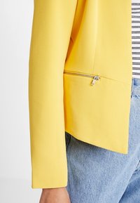 Blazer jaune en tissu texturé, avec une poche zippée sur le côté, porté avec une chemise rayée et un jean en denim.