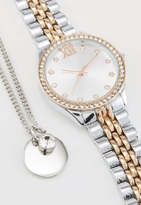 Montre-bracelet métallique avec des tons argent et or rose, cadran rond orné de cristaux, et bracelet à maillons. Accompagnée d'un collier pendentif.