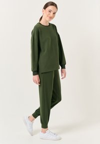 Groene sweatshirt met een ontspannen pasvorm en lange mouwen, gecombineerd met bijpassende tapered joggers. Beide zijn gemaakt van zachte, gestructureerde stof. Witte sneakers.