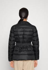 Černá péřová bunda s páskem v pase, s prošívaným designem a širokým límcem. Materiál vypadá hladce a leskle. Pohled z vzadu.