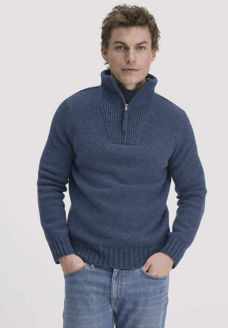 Blauer Strickpullover mit hohem, geripptem Kragen und Reißverschluss. Verfügt über eine strukturierten Passe, enge Bündchen und einen gerippten Saum, getragen über blauen Jeans.