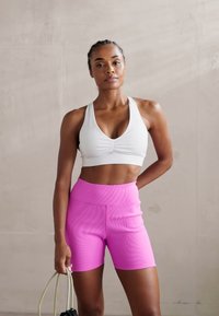Hvit racerback sports-BH med teksturert front, kombinert med høy-midje, glatte rosa sykkelshorts. Begge plaggene har en tettsittende design.