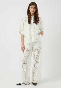 Completo in pizzo bianco a due pezzi, composto da una blusa ampia a maniche corte e pantaloni larghi con motivi floreali e bordi a scallop.