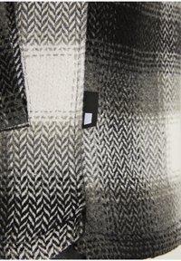Gros plan sur un tissu en laine à chevrons noirs et blancs et à motif écossais, avec une petite étiquette noire cousue dans la couture.