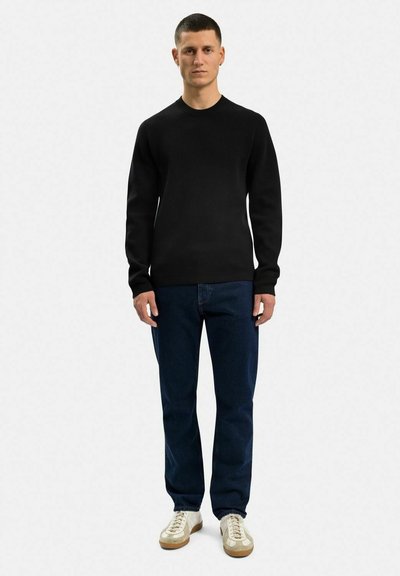 Sort strikket crewneck sweater, parret med mørkeblå denimjeans. Modellen bærer hvide sneakers med beige accenter. Enkel, klassisk design.