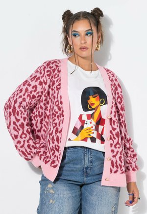 Vrouw met twee space buns kapsel, blauwe oogschaduw, grote oorringen, roze cardigan met luipaardprint, grafisch wit t-shirt en gescheurde spijkerbroek.