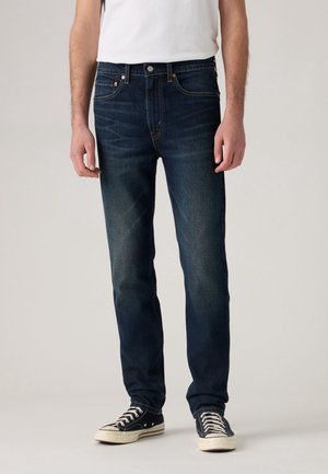 Mężczyzna ubrany w ciemnoniebieskie dżinsy, białą koszulę i czarne trampki, stojący na jednolitym tle.